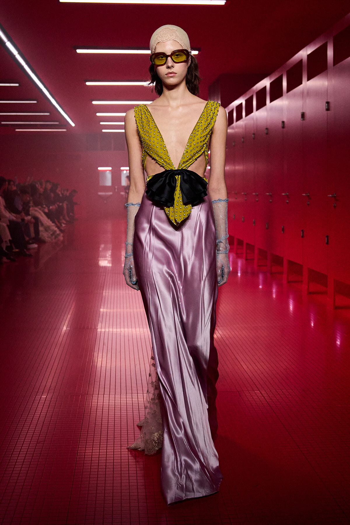 Model on a fashion runway wearing a mustard-yellow studded plunging top, a black bow belt, and lavender satin skirt with sheer gloves, under pink lighting.