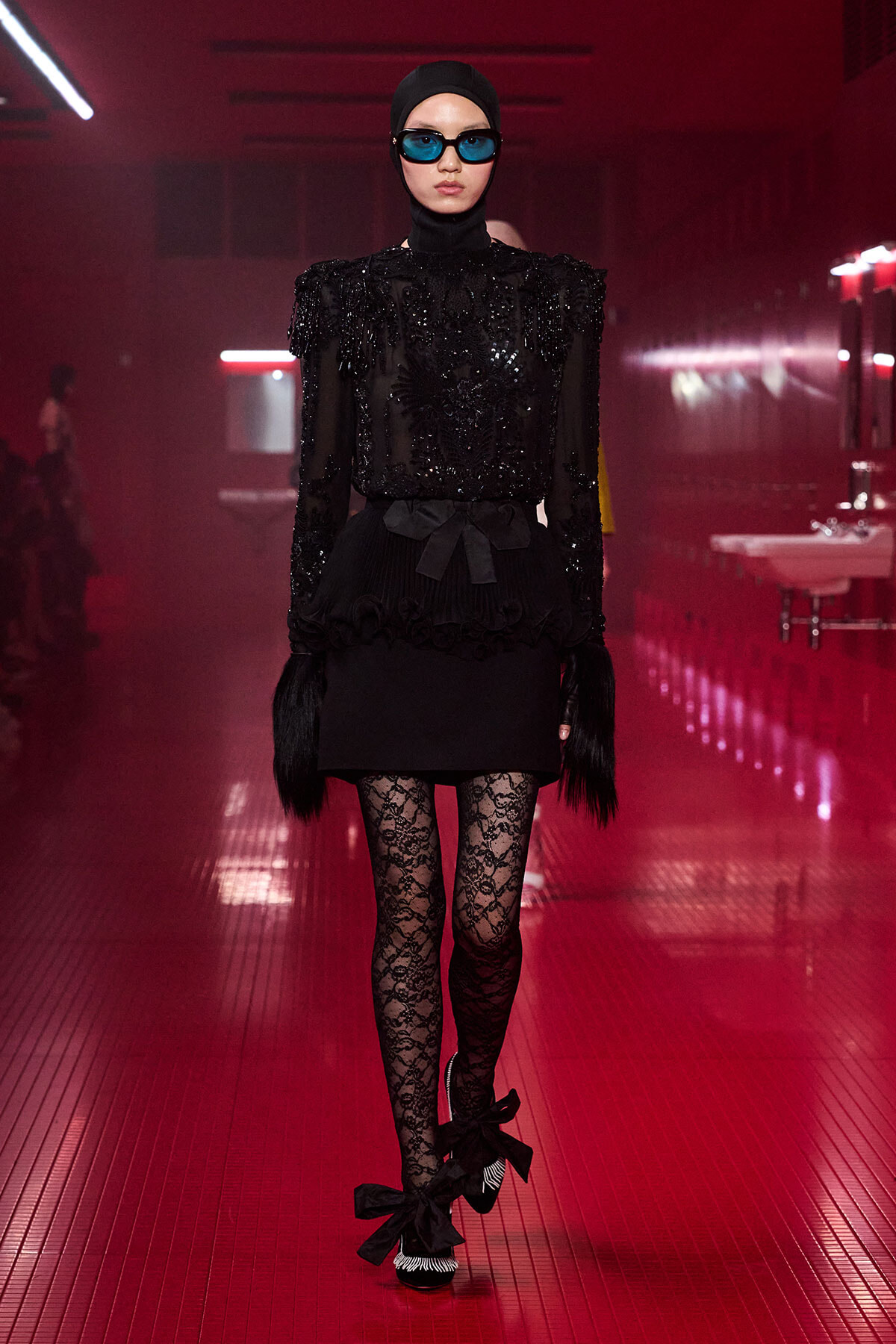 Model on a red-lit runway wearing an all-black, embellished outfit with a hijab and blue-tinted sunglasses. Bold, textured top with a belted waist and lace tights complete the look.