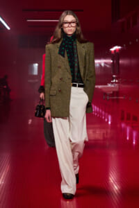 Model wearing an olive tweed blazer over a black turtleneck with a green polka-dot scarf, white wide-leg trousers, and glasses on a red-lit runway.