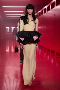Model walking a pink-lit backstage hallway in a pale yellow one-shoulder gown with a large black-and-pink ruffled peplum.