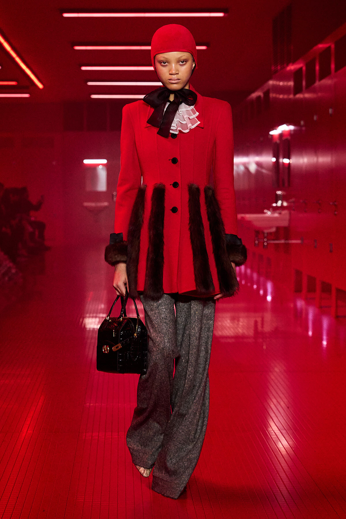 Model on a red-lit runway wearing a bright red coat with fur trim, grey wide-leg pants, and a red cap, carrying a black handbag.