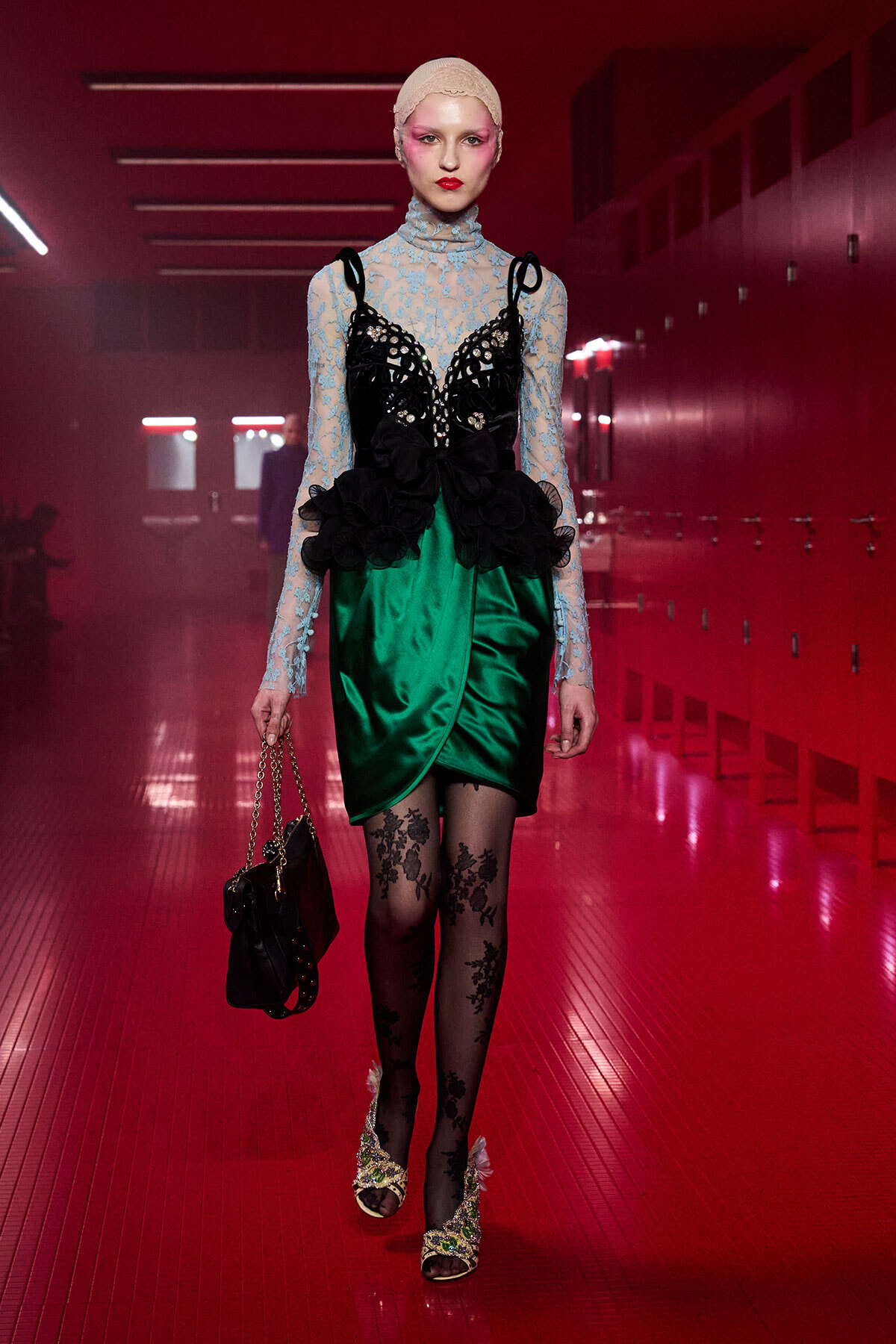 Model walking a runway in a red-lit hallway wearing a blue lace turtleneck, black lace bodice, emerald green satin wrap skirt, and floral tights with embellished heels; carrying a chain-strap bag.