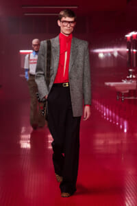 Male model on a red-lit runway wearing a grey blazer, red blouse with bow, black wide-leg trousers, leopard-print shoes, and a patterned bag strapped over the shoulder.