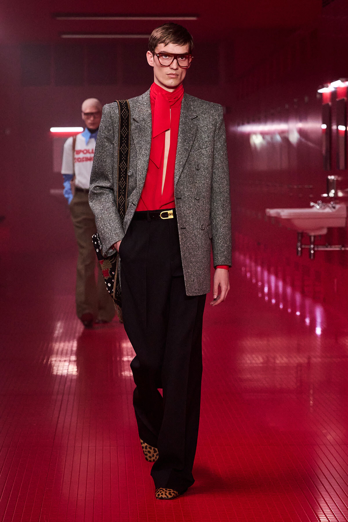 Male model on a red-lit runway wearing a grey blazer, red blouse with bow, black wide-leg trousers, leopard-print shoes, and a patterned bag strapped over the shoulder.
