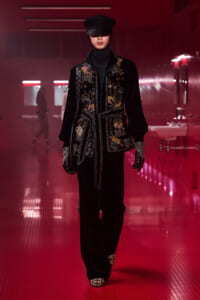 Model walking a runway in a black velvet embroidered jacket with floral appliqués, belted at the waist, paired with a cap, gloves, and dark trousers in a red-lit corridor.