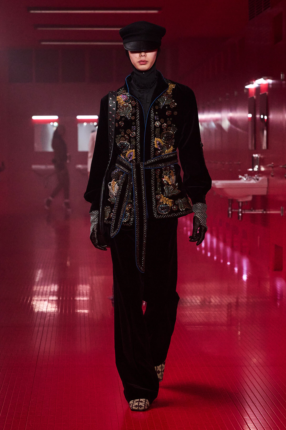 Model walking a runway in a black velvet embroidered jacket with floral appliqués, belted at the waist, paired with a cap, gloves, and dark trousers in a red-lit corridor.