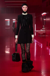 Runway model in a black coat dress with white cuffs and a large black bow, lace tights, fur-trimmed boots, and a black handbag.