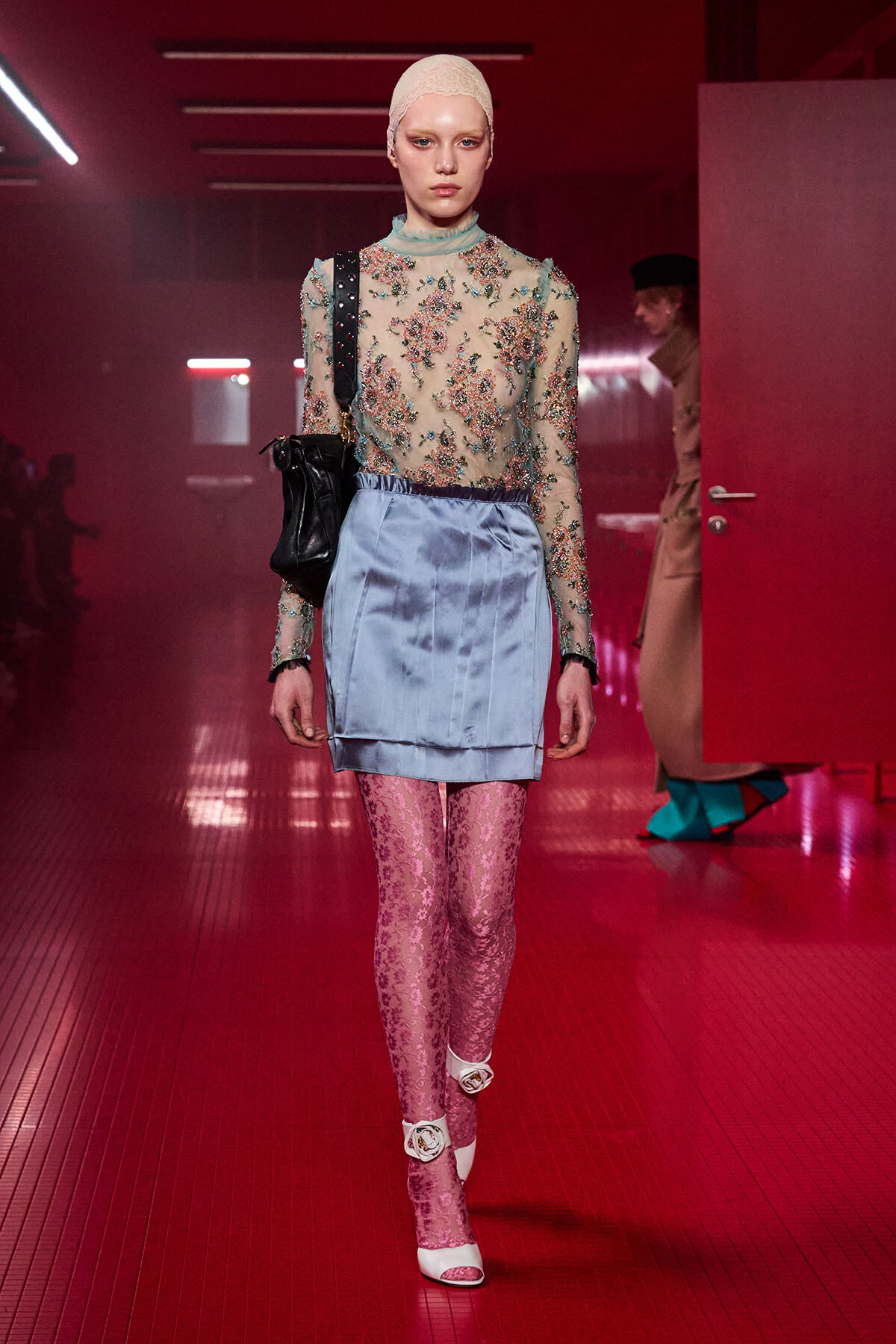Model walking a runway in a sheer embroidered top, pale blue satin mini-skirt, pink lace tights, white heels, and a pale head covering.