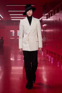 Model on a runway in a white double-breasted blazer, black turtleneck, wide-leg pants, and a black hat, beneath red lighting.