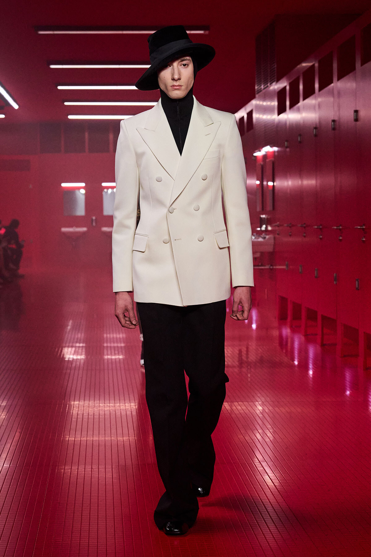 Model on a runway in a white double-breasted blazer, black turtleneck, wide-leg pants, and a black hat, beneath red lighting.