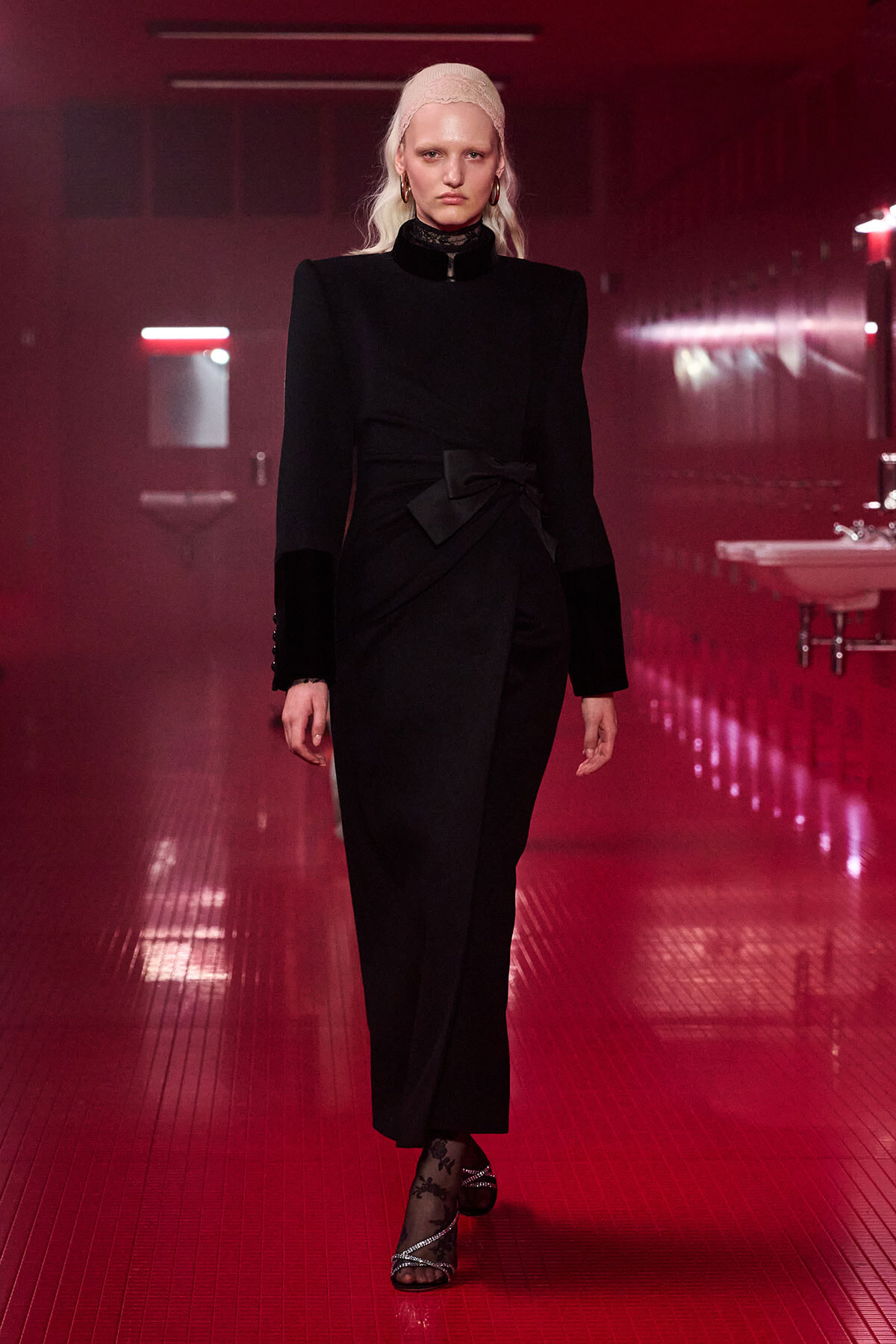 Model walking a runway in a black tailored jumpsuit with a waist bow, in a red-lit hallway.