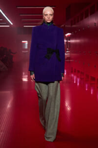 Model walking a fashion runway in a royal purple coat with a black bow, wide gray trousers, and a beige knit cap in a red-lit hallway.