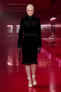 Model walking a red-lit hallway in a black vintage-inspired dress with lace collar, gloves, and white heels, holding a small bag.