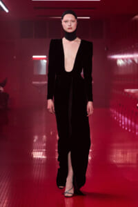 Model in a black velvet jumpsuit with a deep plunging neckline walks a red-lit fashion show runway, wearing a matching head wrap.