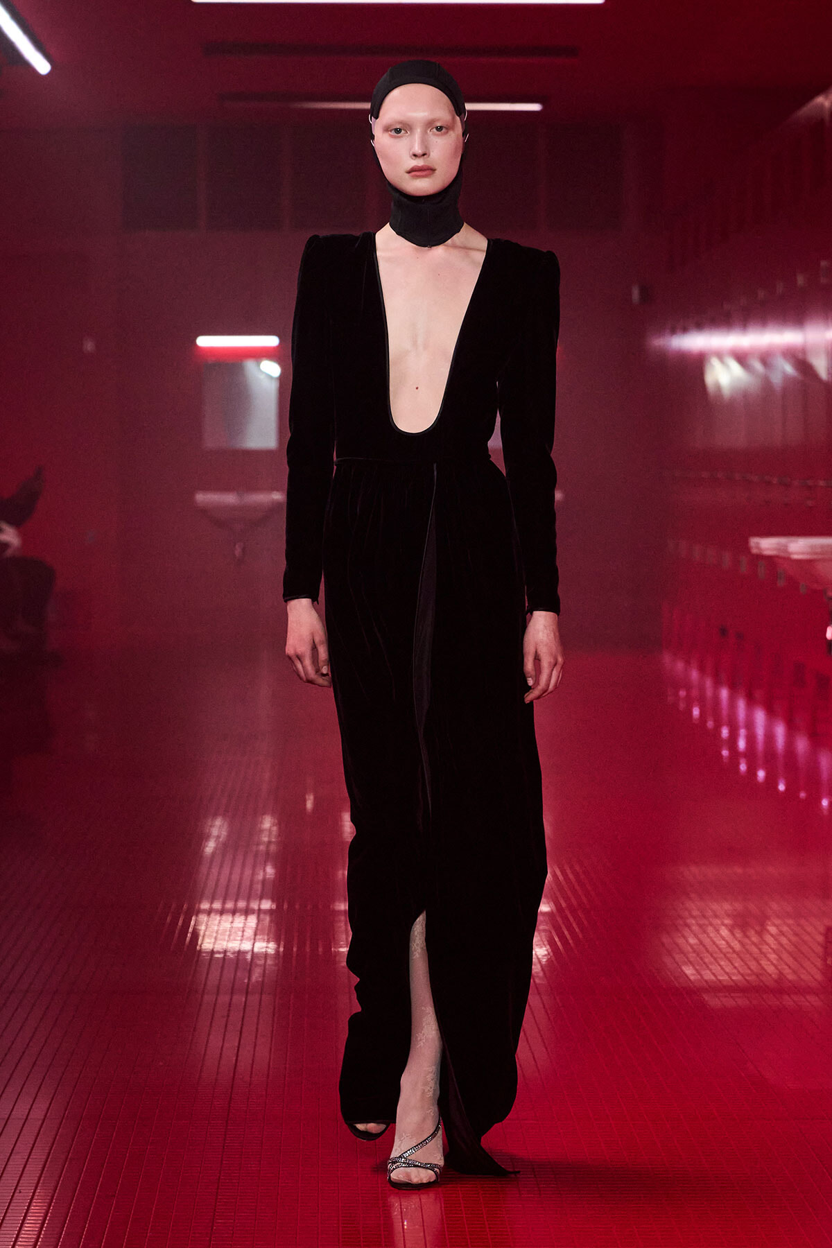 Model in a black velvet jumpsuit with a deep plunging neckline walks a red-lit fashion show runway, wearing a matching head wrap.