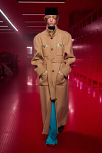 Model walks the runway in a camel belted coat with gold pocket accents, black hat, and bright blue trousers in a red-lit hallway setting