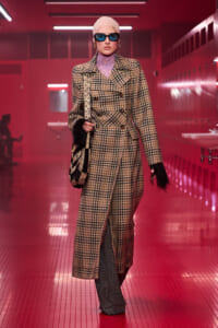 Model walking runway in a beige plaid overcoat, purple lace neck top, and blue sunglasses in a pink-lit corridor with a patterned bag on her shoulder.
