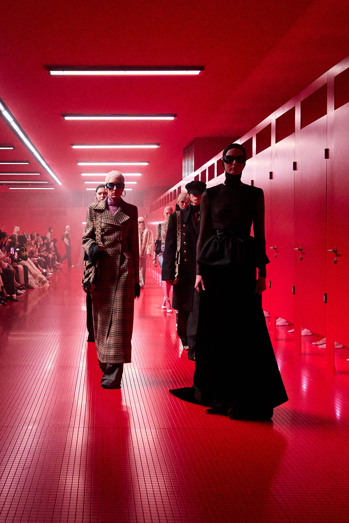 Models walk a red-lit runway in chic outfits during a fashion show.
