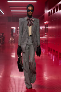 Model walks a runway in a grey double-breasted blazer, matching wide-leg trousers, beige top, pink striped bow, black hijab and sunglasses, carrying a black handbag with chain.