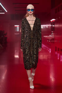 Model walks the runway in a black gold sequined midi dress with a deep V, cut-out strap details, and snake-skin pattern, paired with blue sunglasses and white heels.