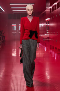 Model walking a red-lit runway in a red blazer with a black bow, gray wide-leg trousers, lace turtleneck, and a pale headband, carrying a small handbag.