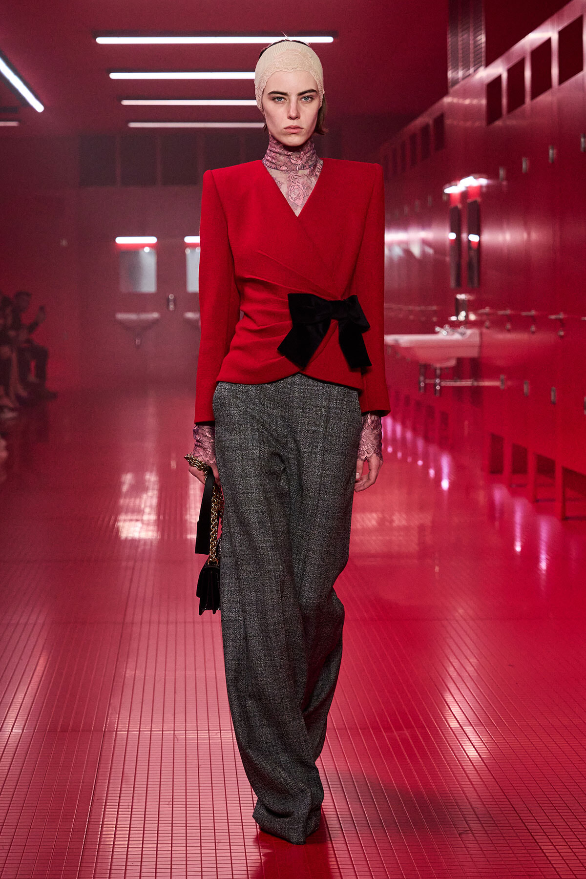 Model walking a red-lit runway in a red blazer with a black bow, gray wide-leg trousers, lace turtleneck, and a pale headband, carrying a small handbag.