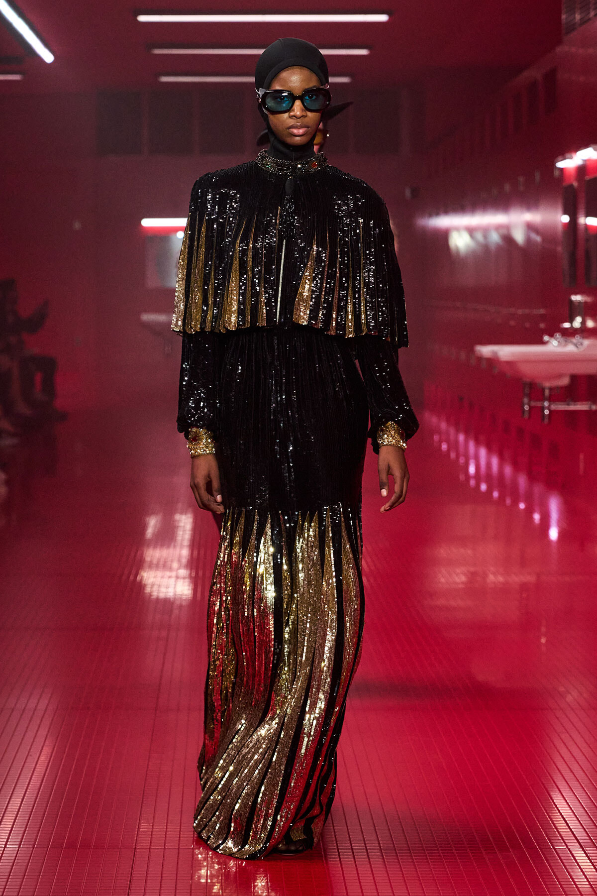 Model walking a runway in a black sequined gown with gold fringe cape, matching cuffs, head wrap, and sunglasses under red lighting.