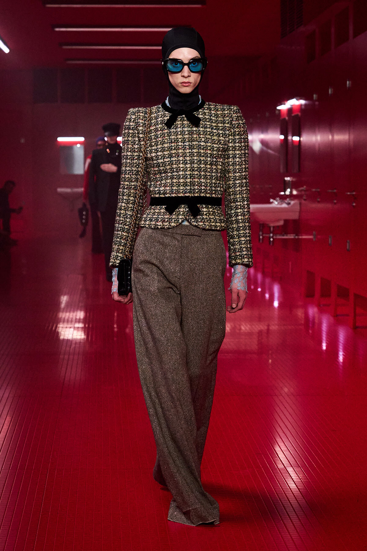 Model walking a runway in a red-lit corridor, wearing a tweed cropped jacket with black bow, wide brown trousers, black headscarf, and blue goggles, carrying a small black bag on a chain.