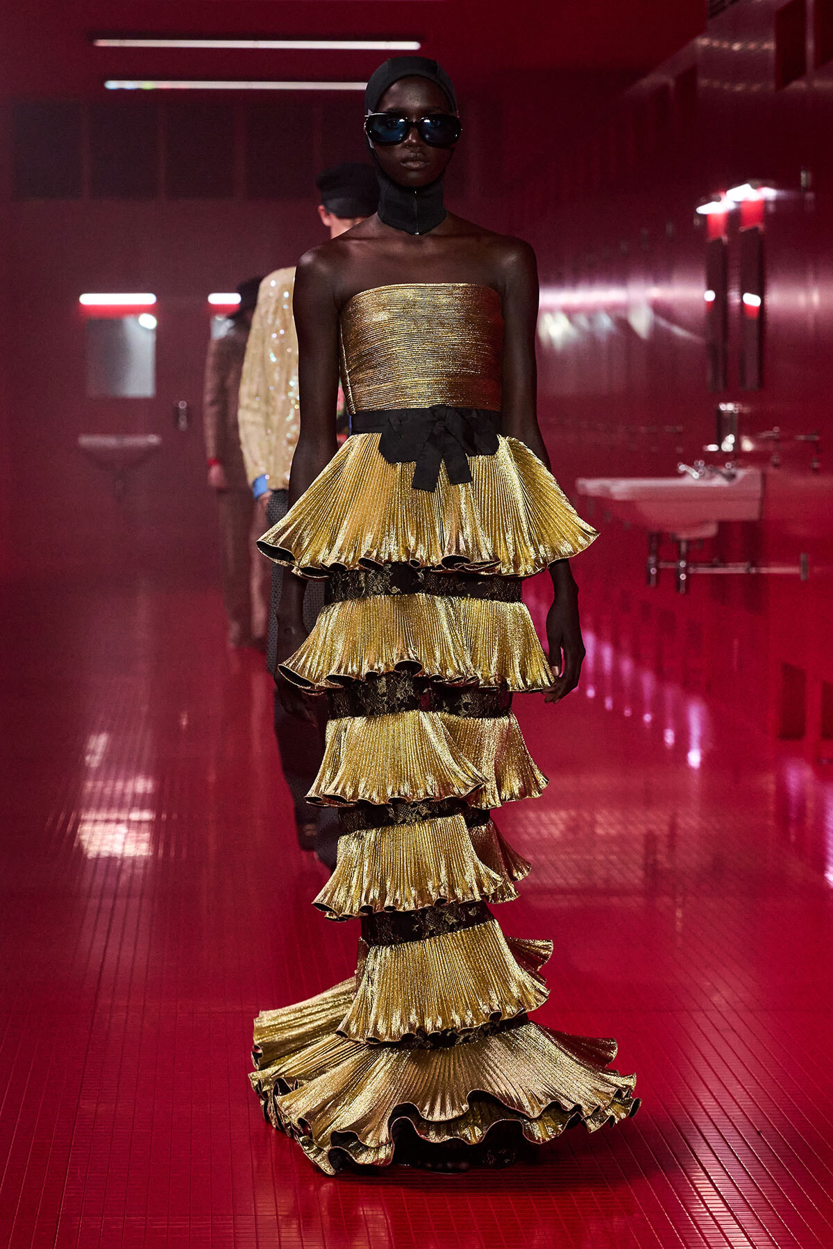 Model on a runway wearing a metallic gold strapless dress with multiple tiered ruffles and a black bow belt, in a red-lit backstage hallway with sunglasses and a head covering.
