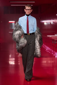 Male model walks down a red-lit hallway in a light blue dress shirt, red patterned tie, gray trousers, brown belt, and a large faux fur coat draped over his shoulders, wearing glasses.