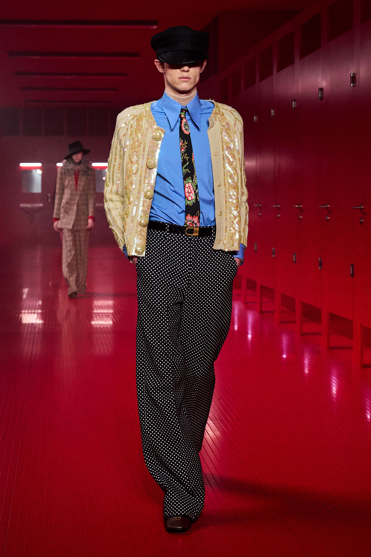 Runway model in a blue shirt, floral tie, cream sequined cardigan, and black polka-dot trousers on a red-lit locker-room runway.