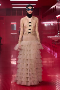 Model in a pink lace gown with a tiered tulle skirt, black ribbon lacing, and blue goggles posing in a pink-tiled hallway with red lighting.