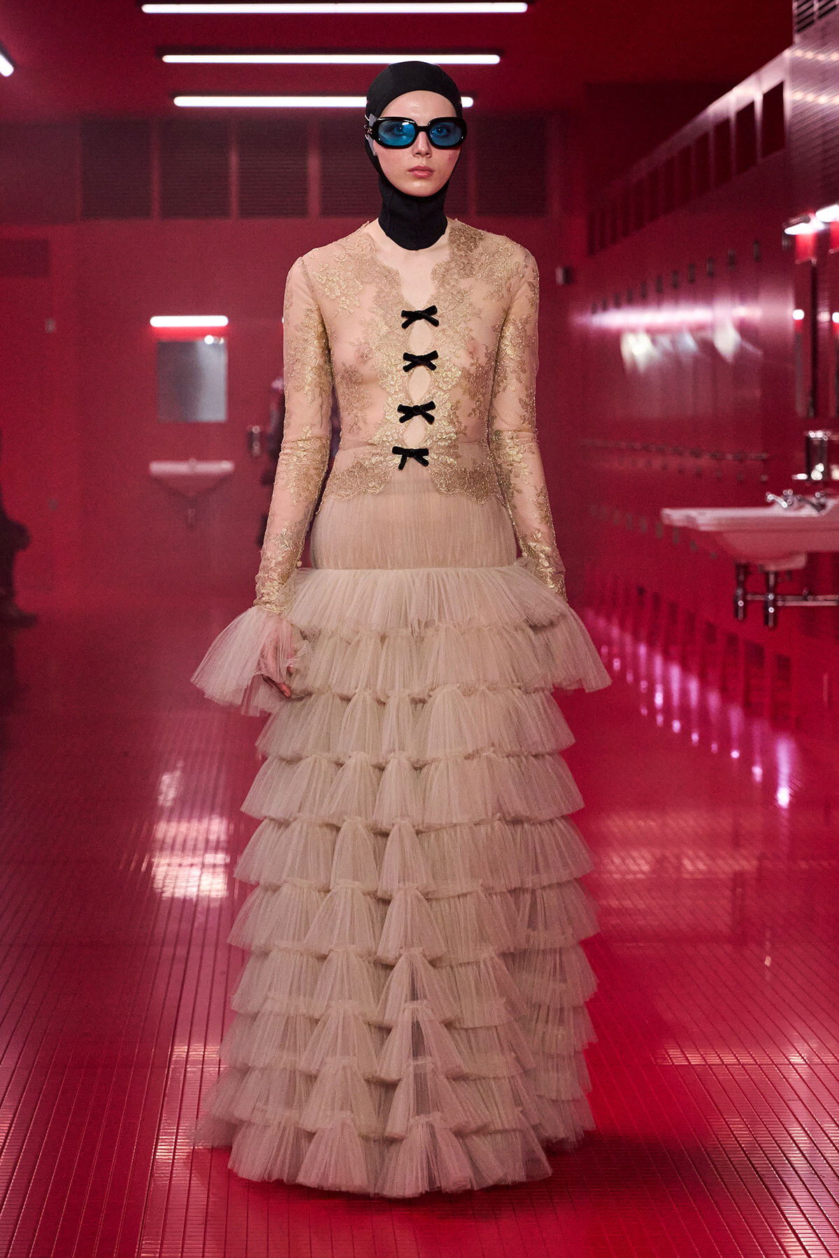 Model in a pink lace gown with a tiered tulle skirt, black ribbon lacing, and blue goggles posing in a pink-tiled hallway with red lighting.