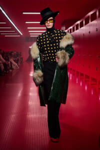 Model walking a fashion show in a black polka-dot blouse with a green fur-trimmed coat, wide-brim hat, and yellow-tinted glasses in a red-lit hallway.
