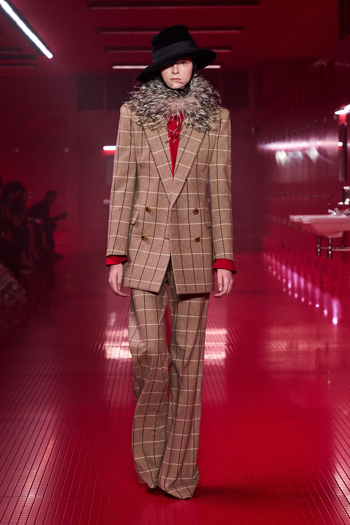 Model on a red-lit runway wearing a beige plaid double-breasted suit, red top, fur collar, and a black wide-brim hat.