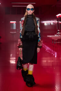 Model on a pink-lit runway wearing a jeweled fur-trimmed jacket over black attire, blue sunglasses, beige cap, and yellow socks with feathered boots, carrying a black handbag.
