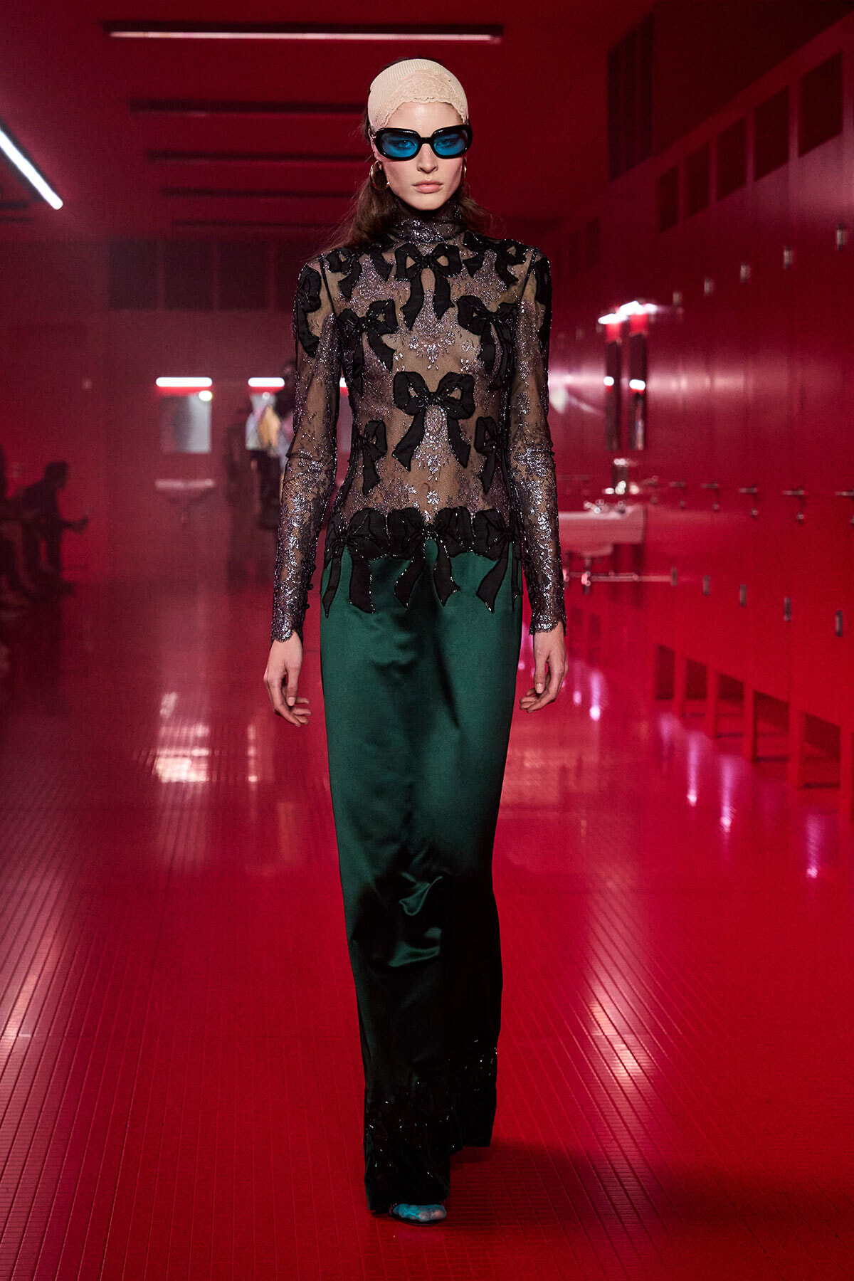 Model walks a fashion show in a red-lit hallway, wearing a sheer black lace blouse with bow motifs and emerald green satin pants, blue sunglasses, and a beige headband.