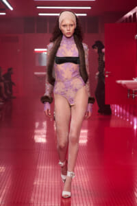 Model walking the runway in a lavender lace bodysuit with fur stole, headband, and pink-lit backstage setting