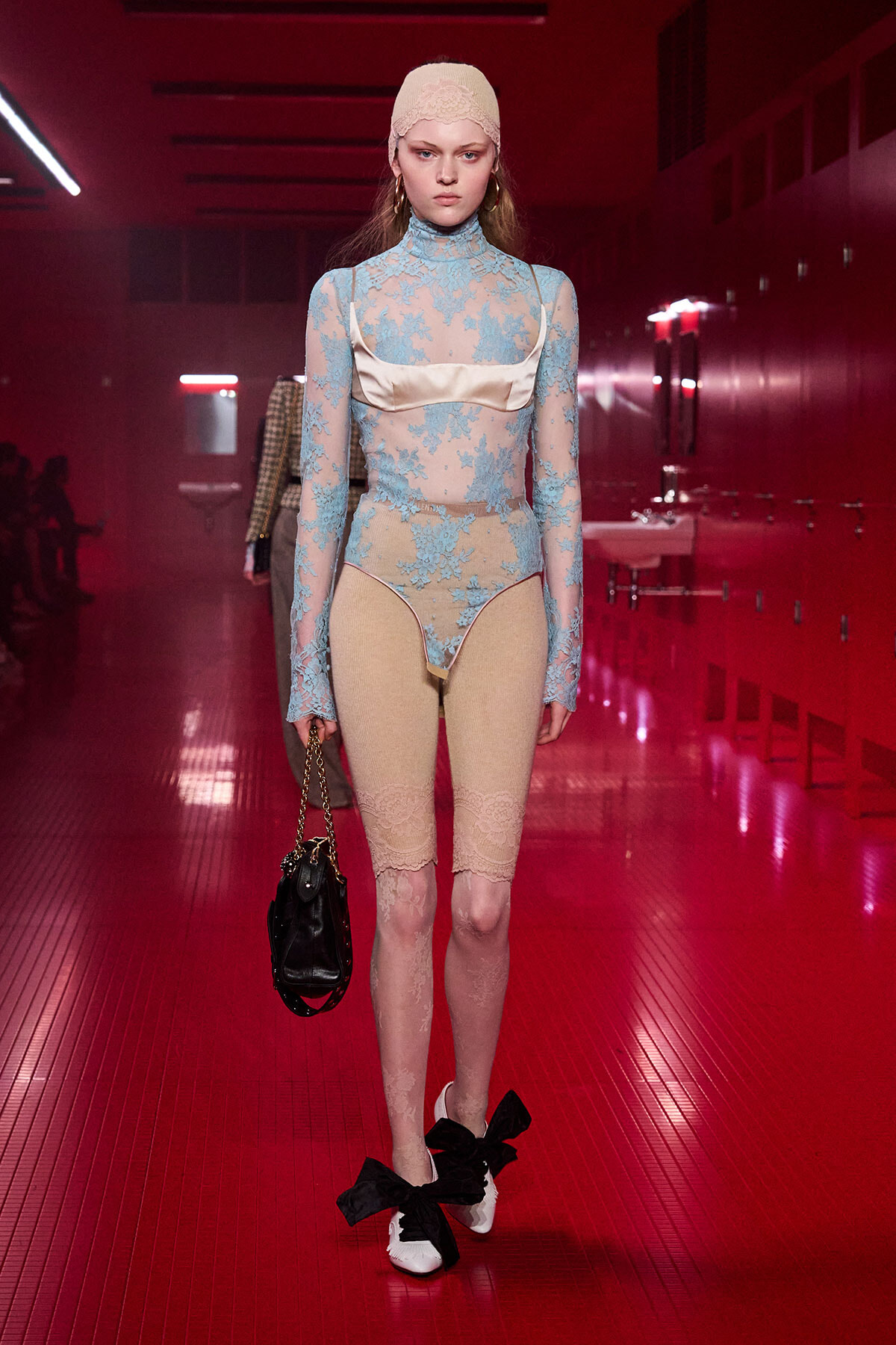 Model walking a runway in a sheer blue lace bodysuit with beige undergarment panels, white shoes with large black bows, and a beige headscarf, carrying a small black handbag.