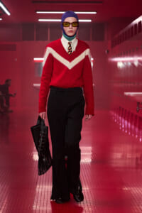 Model walking a runway in a red sweater with a white chevron, white shirt and tie, blue cap, yellow sunglasses, and a black fringed handbag.
