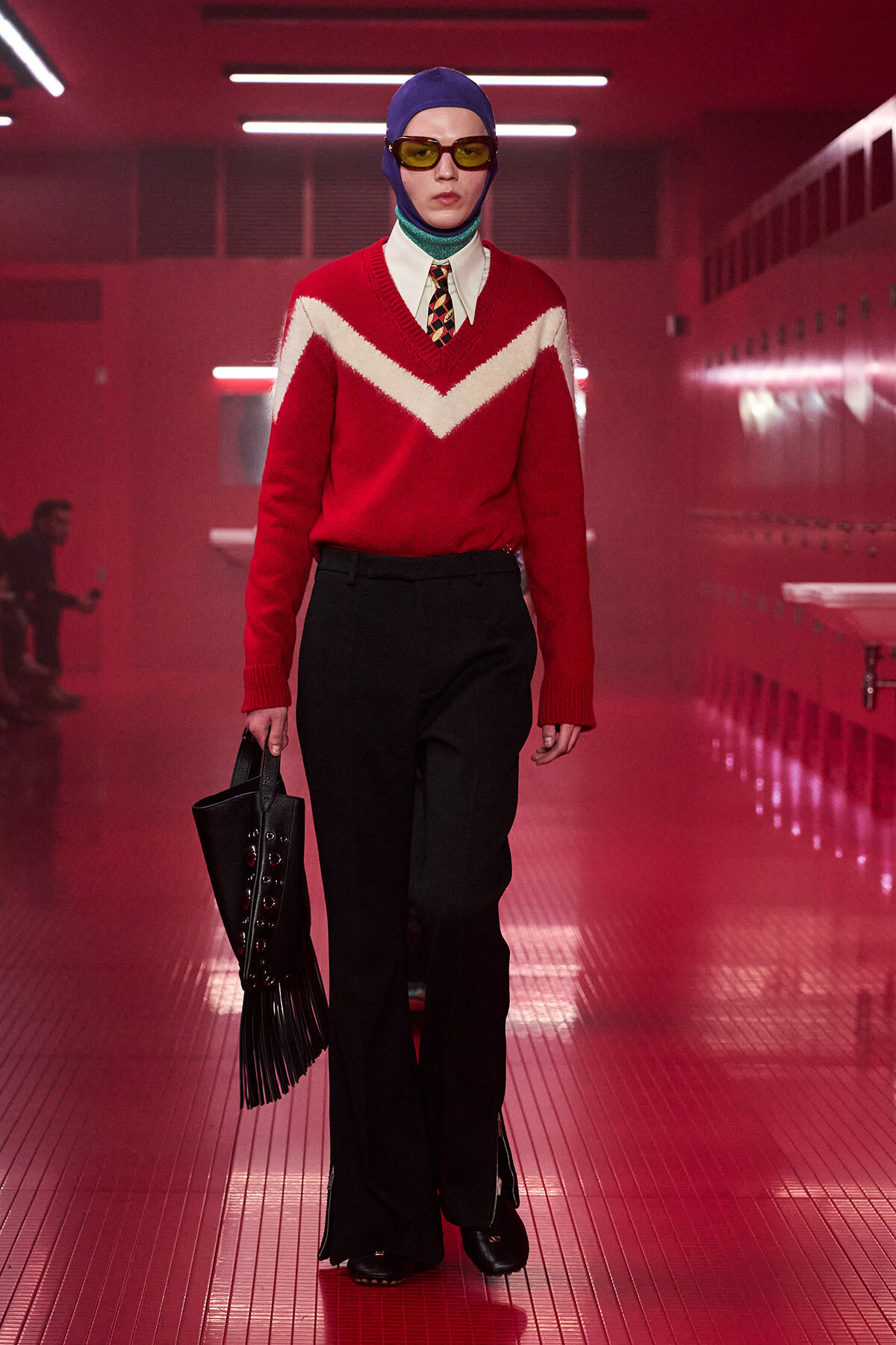 Model walking a runway in a red sweater with a white chevron, white shirt and tie, blue cap, yellow sunglasses, and a black fringed handbag.