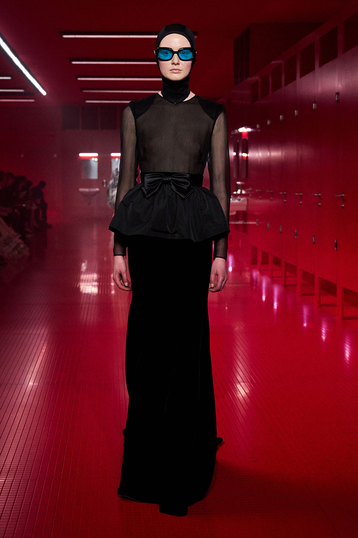 Runway model wearing a sheer black top with a peplum bow and long black skirt, blue goggles, and a black cap in a red-lit hallway.