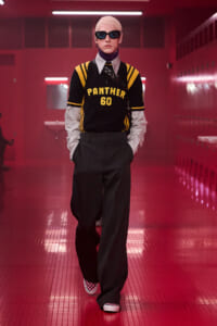 Model walking in a black and yellow Panthers jersey (#60) with oversized dark trousers, pink hair, and sunglasses in a red-lit locker room.