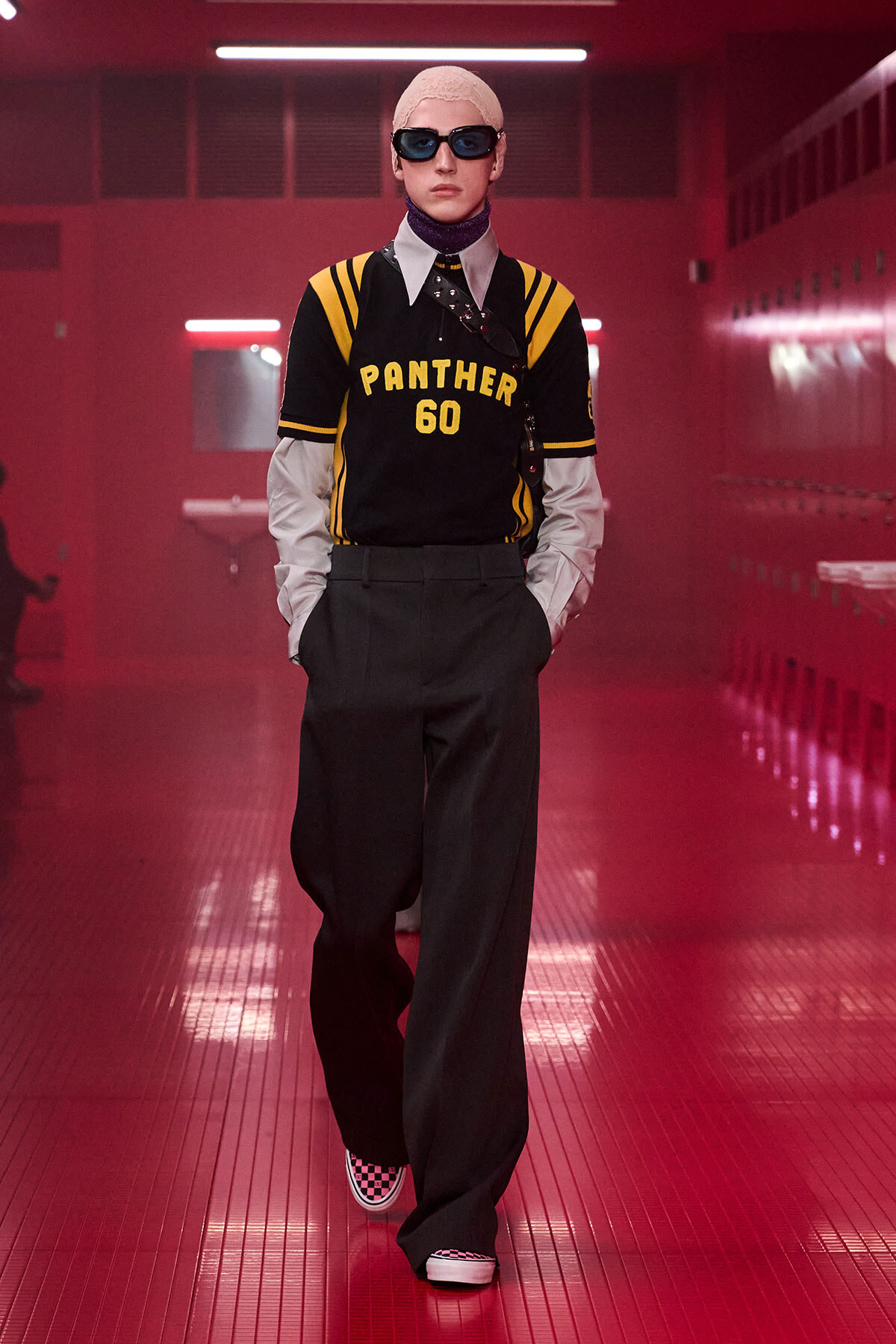 Model walking in a black and yellow Panthers jersey (#60) with oversized dark trousers, pink hair, and sunglasses in a red-lit locker room.