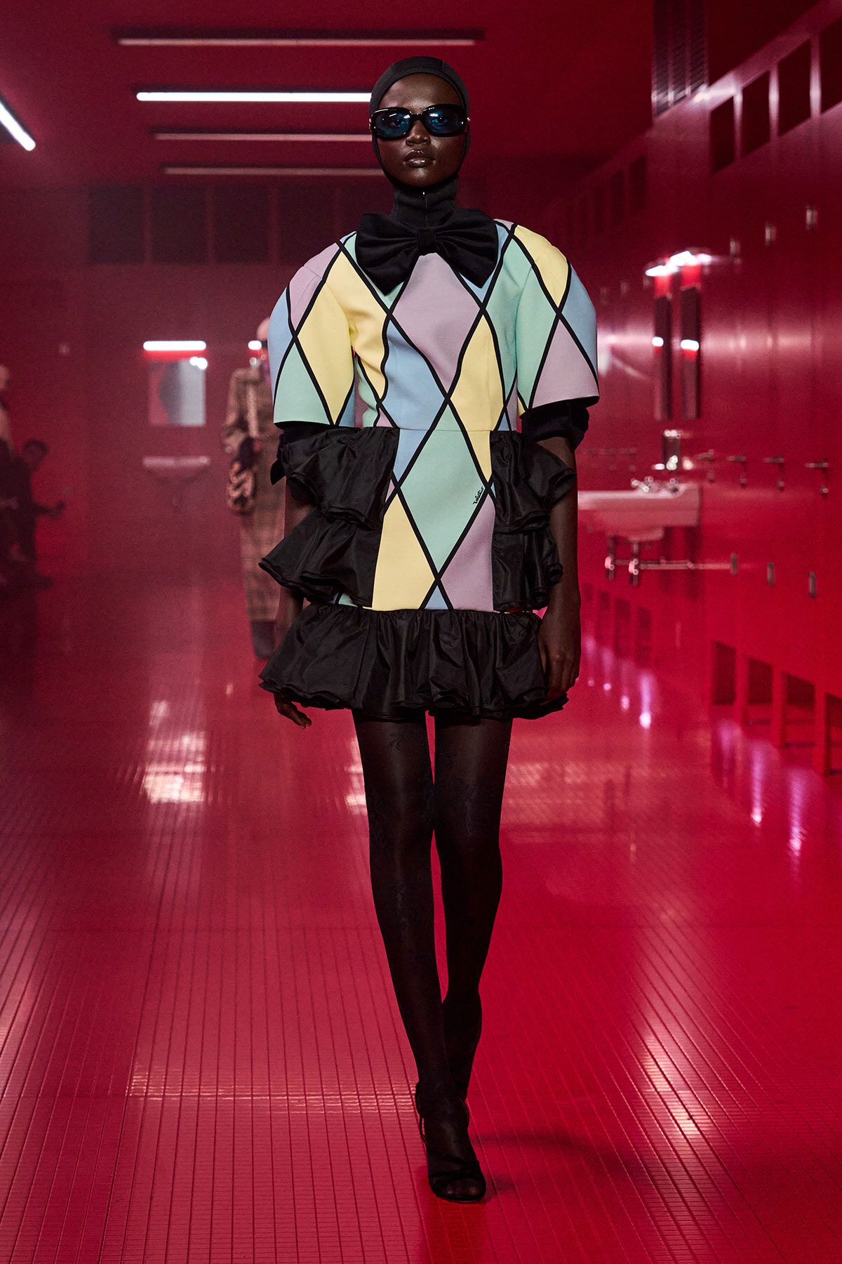Model walking runway in pastel argyle dress with oversized sleeves, black ruffles, and bow tie under red lighting. She wears a head wrap, sunglasses, and black tights.