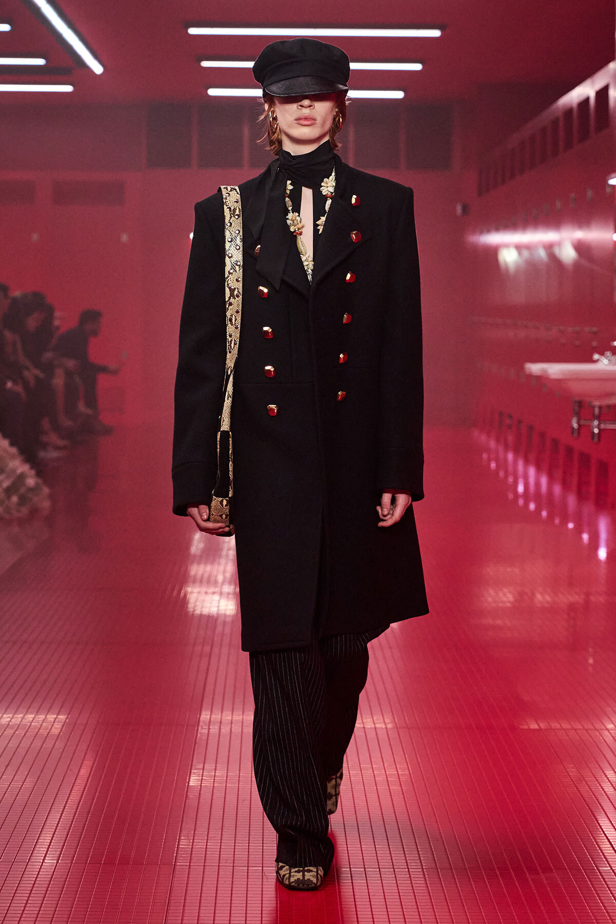 Model walks runway in a black double-breasted coat with red buttons, black cap, striped pants, and a gold-patterned bag strap under red lighting.