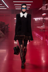 Model walking a red-lit runway in a black coat with a white lace collar, fur trim at the hem, a black headscarf, blue sunglasses, and floral tights while carrying a chain-strap bag.