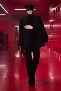 Model walking a red-lit hallway runway wearing a black oversized suit, cap, turtleneck, and beaded necklace.