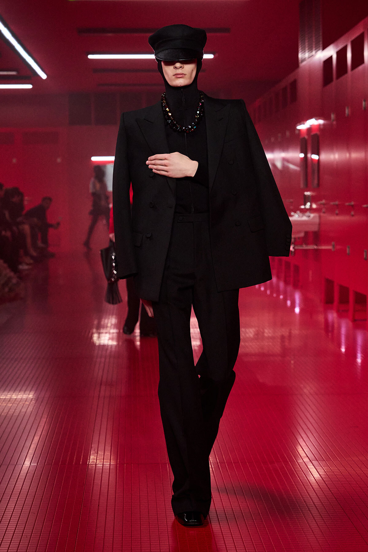 Model walking a red-lit hallway runway wearing a black oversized suit, cap, turtleneck, and beaded necklace.