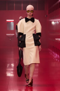 Model walking a runway in a pale pink dress with black peplum ruffles, oversized bow, and sunglasses, carrying a black studded bag.